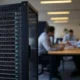 A modern office meeting with professionals discussing around a table, featuring a large server rack in the foreground representing legacy systems integrated with contemporary technology, ideal for finance and healthcare industries.
